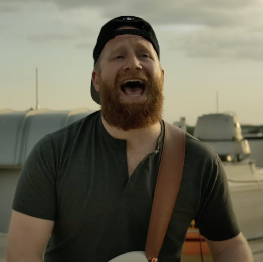 Bearded man in a backwards cap singing outdoors with a guitar strap, moored boats behind